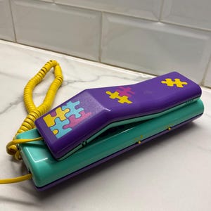 Può includere: Un telefono vintage con una cornetta viola con disegni di puzzle gialli, rosa e blu. La base è verde acqua e il cavo a spirale è giallo. Il telefono è appoggiato su una superficie bianca.