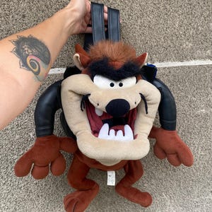 Może przedstawiać: Pluszowy plecak z Tazmanian Devil w czarnej skórzanej kurtce, brązowym futrem i szerokim uśmiechem. Plecak ma regulowane paski.