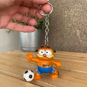 Könnte beinhalten: Orangener Garfield-Schlüsselanhänger mit blauen Shorts und einem Fußball. Der Schlüsselanhänger hat eine silberne Kette und einen Ring. Das Wort "GOAL!" ist auf der Vorderseite von Garfield's Shirt aufgedruckt. Der Fußball ist weiß mit schwarzen Fünfecken.