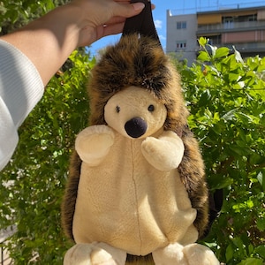 May include: A plush hedgehog backpack with a brown and tan color scheme. The backpack features a soft, stuffed body with a detailed face, including a black nose and small eyes. The backpack has a brown strap for carrying.
