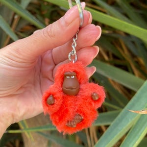 May include: A bright orange fluffy monkey keyring with brown eyes and a brown face. The monkey is attached to a silver chain and ring.
