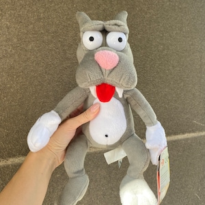 May include: Grey plush toy dog with large white eyes, a pink nose, and a red tongue sticking out. The toy has white paws and is sitting on a brown surface.