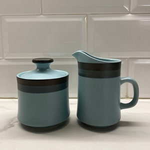 Op de afbeelding: Een blauwe en bruine keramische roomkan en suikerpot set. De roomkan heeft een handvat en de suikerpot heeft een deksel.