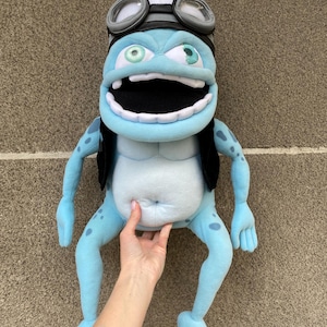 Può includere: Un peluche di un personaggio di rana dei cartoni animati, principalmente azzurro con pancia bianca. La rana ha grandi occhi espressivi, bocca spalancata e indossa un casco bianco con occhiali. Ha un gilet nero e macchie blu.
