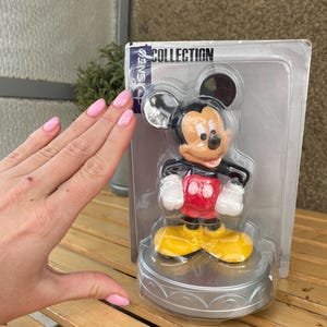 May include: A plastic figurine of Mickey Mouse in a red shirt and yellow shoes. The figurine is on a silver base and is packaged in a clear plastic box with the words "Disney Collection" printed on the top.