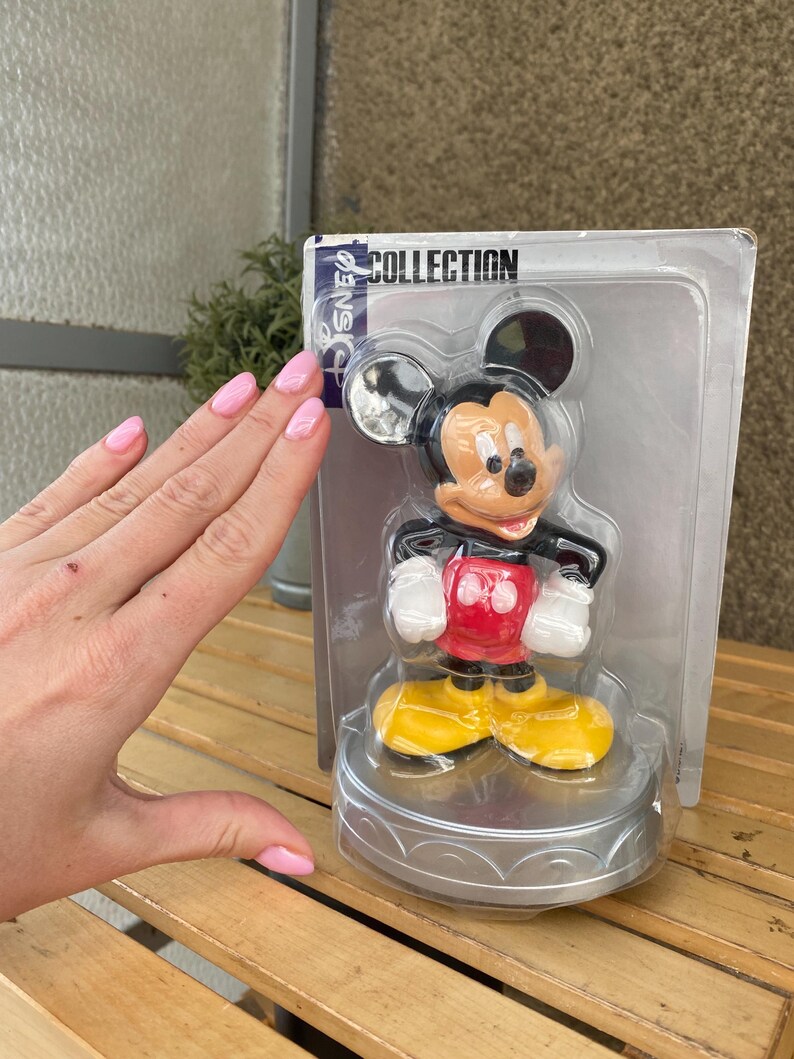 May include: A plastic figurine of Mickey Mouse in a red shirt and yellow shoes. The figurine is on a silver base and is packaged in a clear plastic box with the words "Disney Collection" printed on the top.