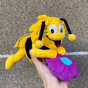 Könnte beinhalten: Ein gelb-schwarzes Plüschspielzeug in Form einer Biene mit großen Augen und einem roten, lächelnden Mund. Die Biene hält ein lila Kissen mit einem türkisfarbenen Design. Ein farbenfrohes Kinderspielzeug.