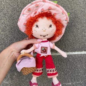 May include: A plush doll with bright orange yarn hair, wearing a pink strawberry-patterned sun hat, a light pink t-shirt with a strawberry graphic, red trousers with a strawberry trim, and pink sandals, while holding a small woven basket.