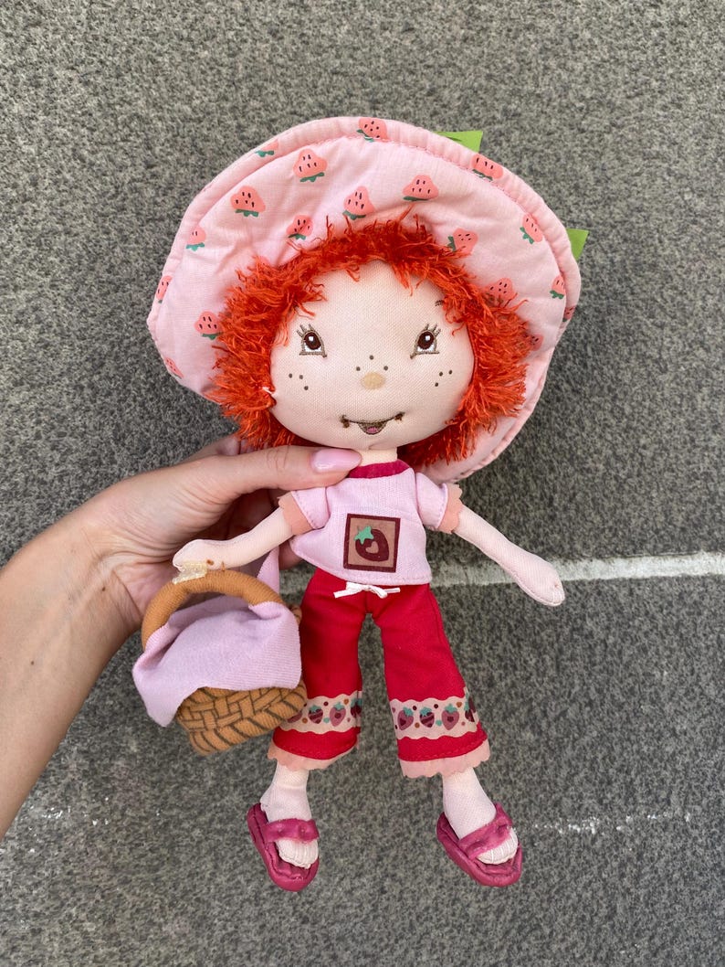 May include: A plush doll with bright orange yarn hair, wearing a pink strawberry-patterned sun hat, a light pink t-shirt with a strawberry graphic, red trousers with a strawberry trim, and pink sandals, while holding a small woven basket.
