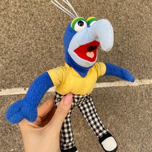 May include: A plush toy of the character Gonzo with blue skin, a long curved nose, and three white hair tufts. The figure wears a yellow short-sleeved shirt, black and white checkered pants, and black shoes with white toe caps, featuring a red mouth.