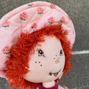 May include: A plush doll with bright orange yarn hair, wearing a light pink fabric hat featuring a repeating strawberry pattern. The doll has embroidered facial features, including brown eyes, freckles, and a small mouth, and wears a pink shirt.
