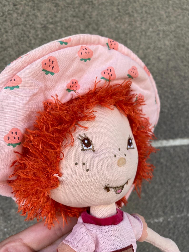 May include: A plush doll with bright orange yarn hair, wearing a light pink fabric hat featuring a repeating strawberry pattern. The doll has embroidered facial features, including brown eyes, freckles, and a small mouth, and wears a pink shirt.