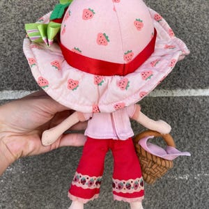 May include: A plush doll wearing a light pink sun hat with a strawberry print, a red ribbon band, and a green bow. The doll has a light pink short-sleeved top, red cropped trousers with a strawberry pattern at the hem, and red shoes, carrying a brown basket.