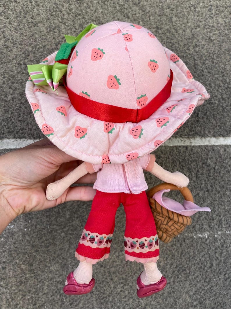 May include: A plush doll wearing a light pink sun hat with a strawberry print, a red ribbon band, and a green bow. The doll has a light pink short-sleeved top, red cropped trousers with a strawberry pattern at the hem, and red shoes, carrying a brown basket.