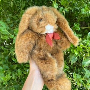 Könnte beinhalten: Ein braunes Plüschkaninchen mit langen Ohren und einer weißen Schnauze. Das Stofftier hat eine rote Schleife um den Hals. Das Kaninchen besteht aus weichem, strukturiertem Material und wird vor grünem Laub gehalten.
