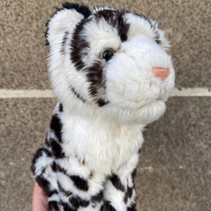 May include: A plush white tiger cub with black stripes and spots. The stuffed animal has a pink nose, black eyes, and a soft, fluffy texture. The toy is designed to resemble a baby tiger.