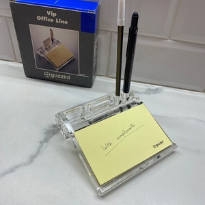 May include: A clear acrylic desk organiser with a notepad and two pens. The notepad has the words "With compliments" written on it. A box labelled "Vip Office Line" is in the background.