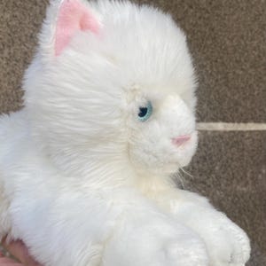 May include: White plush toy cat with pink ears and blue eyes. The cat is lying on its side with its head turned to the right.