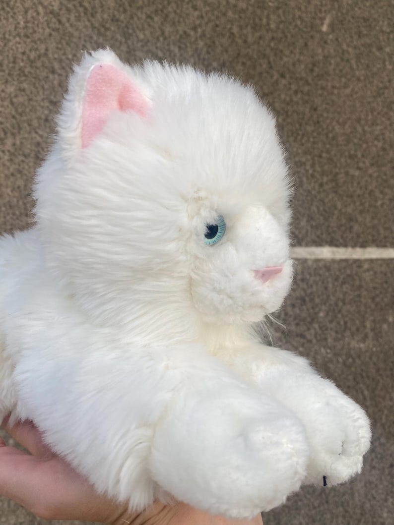 May include: White plush toy cat with pink ears and blue eyes. The cat is lying on its side with its head turned to the right.
