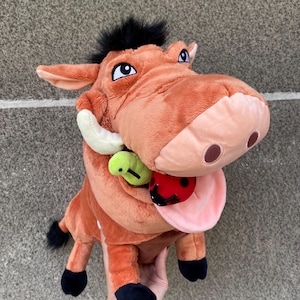 Könnte beinhalten: Ein Plüschtier von Pumbaa aus Der König der Löwen, mit hellbraunem Körper, schwarzer Mähne und Stoßzähnen. Das Spielzeug hat eine rosa Zunge, einen grünen Wurm und einen roten Marienkäfer.