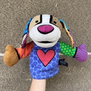 May include: A vibrant dog hand puppet featuring a blue polka-dot shirt with a red heart. The puppet has a tan head, pink nose, and patterned ears and arms. It is a Britto Pop Art design.