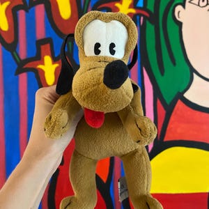 Pode incluir: Um peluche da personagem Pluto, com pelo castanho claro, orelhas pretas compridas, nariz preto e língua vermelha. A personagem tem grandes manchas brancas à volta dos olhos com pupilas pretas e uma textura macia e felpuda em todo o corpo.
