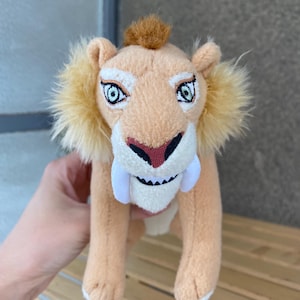 Op de afbeelding: Een pluche sabeltandtijger met lichtbruine vacht, een kleine pluk bruin haar op de kop en pluizige gele wangen. Het speelgoed heeft geborduurde ogen met donkere randen, een donkerrode neus en twee opvallende witte vilten slagtanden.