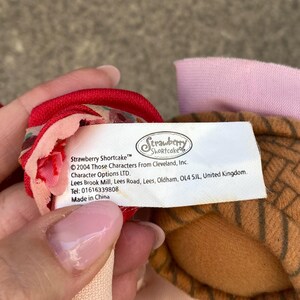 May include: A white fabric product tag with black text reading Strawberry Shortcake, 2004 Those Characters From Cleveland, Inc., Character Options LTD., Lees Brook Mill, Lees Road, Oldham, OL4 5JL, United Kingdom, Tel: 01616339808, Made in China.