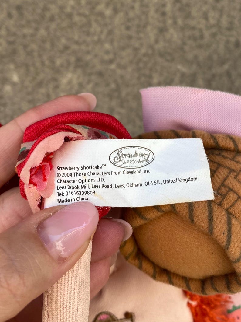May include: A white fabric product tag with black text reading Strawberry Shortcake, 2004 Those Characters From Cleveland, Inc., Character Options LTD., Lees Brook Mill, Lees Road, Oldham, OL4 5JL, United Kingdom, Tel: 01616339808, Made in China.