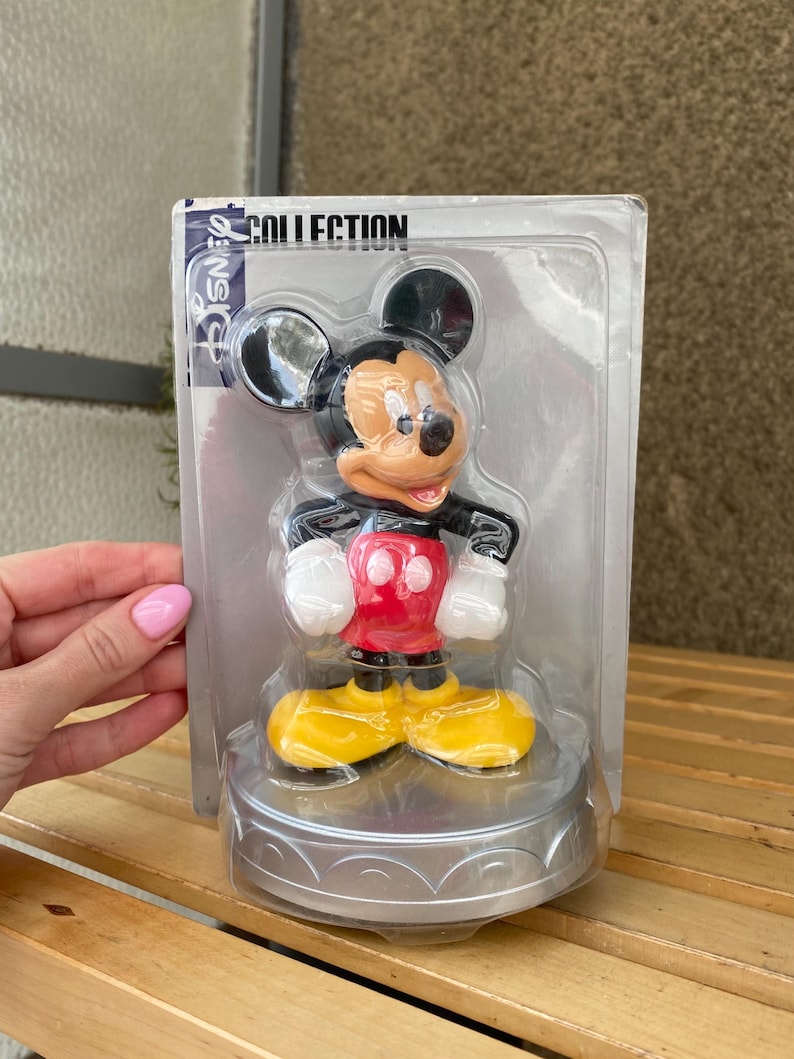 May include: A plastic Mickey Mouse figurine in a clear plastic packaging. The figurine is red, black, and yellow. The packaging has the text "Disney Collection" on it.
