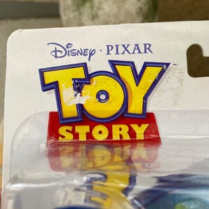 May include: A close-up of a Toy Story packaging with the Disney Pixar logo and the words "Toy Story" in red and blue letters. The packaging is clear plastic and shows a blue and yellow toy inside.
