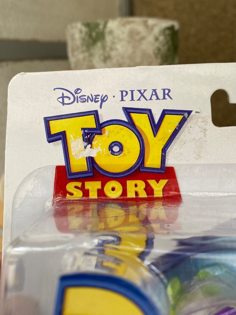 May include: A close-up of a Toy Story packaging with the Disney Pixar logo and the words "Toy Story" in red and blue letters. The packaging is clear plastic and shows a blue and yellow toy inside.