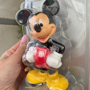 May include: A plastic figurine of Mickey Mouse in a red shirt, yellow shoes, and black shorts. The figurine is packaged in a clear plastic clamshell.
