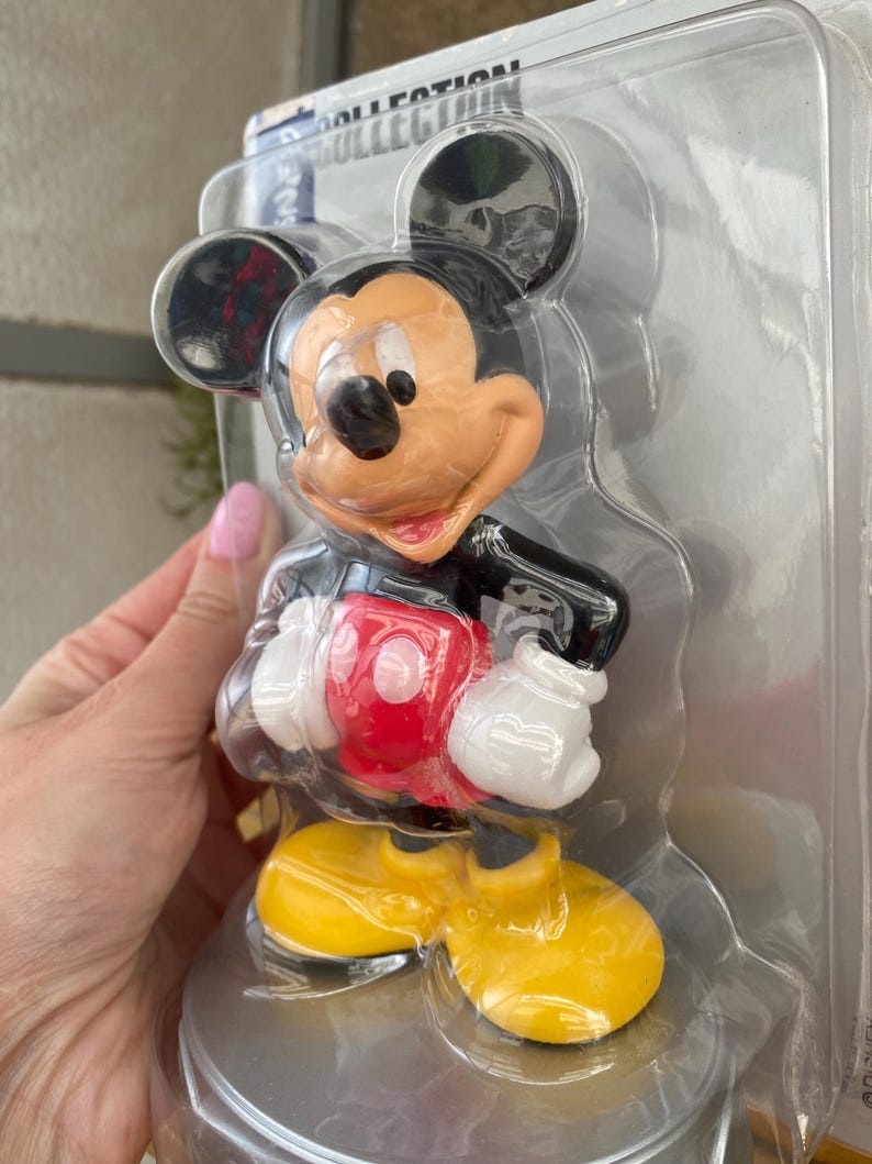 May include: A plastic figurine of Mickey Mouse in a red shirt, yellow shoes, and black shorts. The figurine is packaged in a clear plastic clamshell.