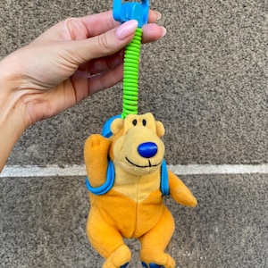 May include: A soft toy bear with orange fur, a blue nose, and a blue backpack. The bear is attached to a blue plastic clip with a green coiled spring. The toy is designed for babies and toddlers.