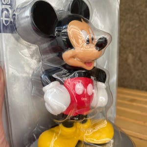 May include: A plastic Mickey Mouse figurine in a clear plastic packaging. Mickey is wearing his signature red shorts with two white buttons, yellow shoes, and white gloves. He is standing on a silver base.