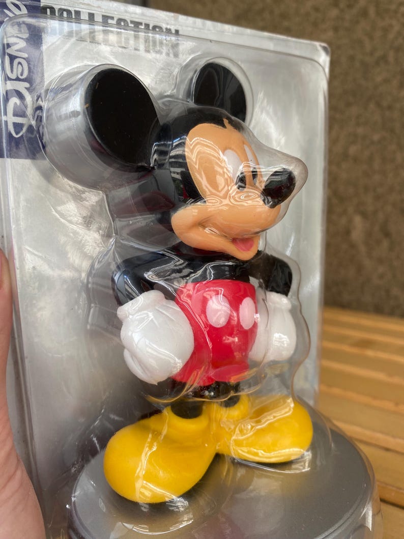 May include: A plastic Mickey Mouse figurine in a clear plastic packaging. Mickey is wearing his signature red shorts with two white buttons, yellow shoes, and white gloves. He is standing on a silver base.