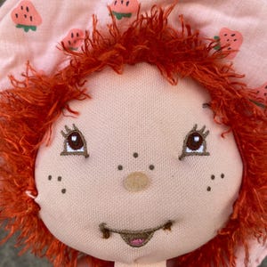 May include: A plush doll with bright orange yarn hair and a light pink bonnet featuring a repeating strawberry pattern. The doll has embroidered brown eyes, a small round nose, freckles, and a stitched mouth with a hint of pink on the lips.
