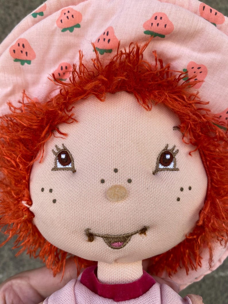 May include: A plush doll with bright orange yarn hair and a light pink bonnet featuring a repeating strawberry pattern. The doll has embroidered brown eyes, a small round nose, freckles, and a stitched mouth with a hint of pink on the lips.