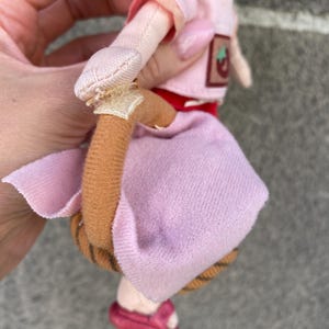 May include: A plush doll with bright orange hair, wearing a light pink short-sleeved top with a small strawberry patch and a light pink skirt. The doll features a brown curved tail attached with a light-coloured fabric tab and dark pink shoes.