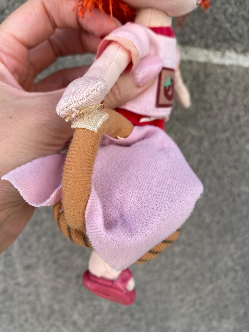 May include: A plush doll with bright orange hair, wearing a light pink short-sleeved top with a small strawberry patch and a light pink skirt. The doll features a brown curved tail attached with a light-coloured fabric tab and dark pink shoes.