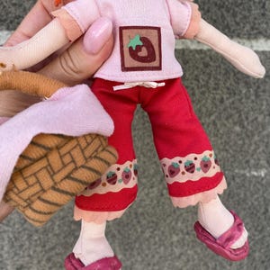 May include: A plush doll with bright orange hair wearing a light pink short-sleeved shirt featuring a brown square patch with a strawberry graphic, red cropped trousers with a strawberry print hem, and pink sandals, while holding a small woven basket.