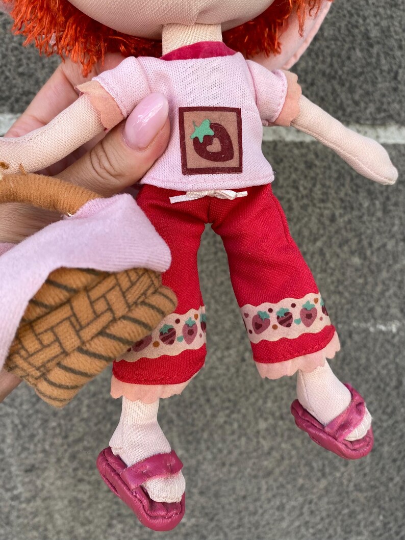 May include: A plush doll with bright orange hair wearing a light pink short-sleeved shirt featuring a brown square patch with a strawberry graphic, red cropped trousers with a strawberry print hem, and pink sandals, while holding a small woven basket.