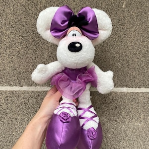 Könnte beinhalten: Ein weißes Plüschmaus-Spielzeug mit einer großen lila Satinschleife, einem Tutu und Ballettschuhen. Die Maus hat eine schwarze Nase und Augen, und die Ballettschuhe haben weiße Bänder und Rosen. Das Spielzeug ist weich und flauschig.