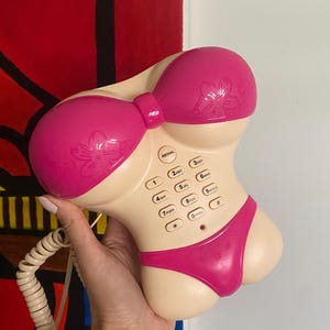 Peut inclure: Un téléphone fantaisie en forme de bikini. Le téléphone est principalement beige avec des accents roses, notamment les bonnets de soutien-gorge et le bas de bikini. Le téléphone possède un clavier standard avec des chiffres et des touches de fonction.