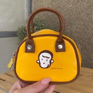 Op de afbeelding: Een kleine, gele handtas met bruine handvatten en randen. De tas heeft een cartoon apengezicht en Japanse tekst. Een bananenvormige ritstrekker voegt een speels tintje toe. De tas is ontworpen met een afgeronde vorm.