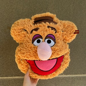 May include: A plush brown and orange puppet with a pink nose, red mouth, and purple eyelids. The puppet is smiling and has a brown stripe across its forehead. The puppet is labelled "Tippets".