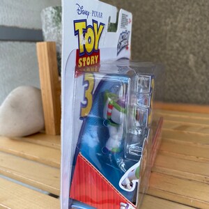 May include: A plastic package containing a Buzz Lightyear action figure from the Disney Pixar Toy Story franchise. The figure is wearing a white and green spacesuit with a red stripe and a blue helmet. The packaging is blue and red with the Toy Story logo and the words "Disney Pixar" printed on it.