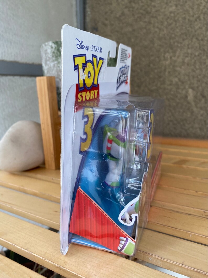 May include: A plastic package containing a Buzz Lightyear action figure from the Disney Pixar Toy Story franchise. The figure is wearing a white and green spacesuit with a red stripe and a blue helmet. The packaging is blue and red with the Toy Story logo and the words "Disney Pixar" printed on it.