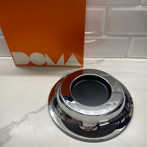 May include: A chrome-plated ashtray with a black interior, reflecting the surrounding environment. The ashtray is circular with a raised center and a wide, flat base. An orange box with the word "DOMA" and the brand "iGuzzini" is in the background.